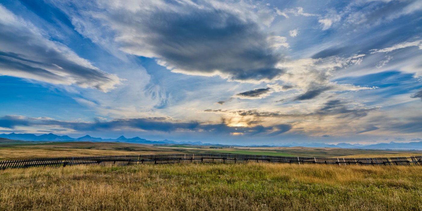 a field with a fence in the foreground and a cloudy sky in the background .