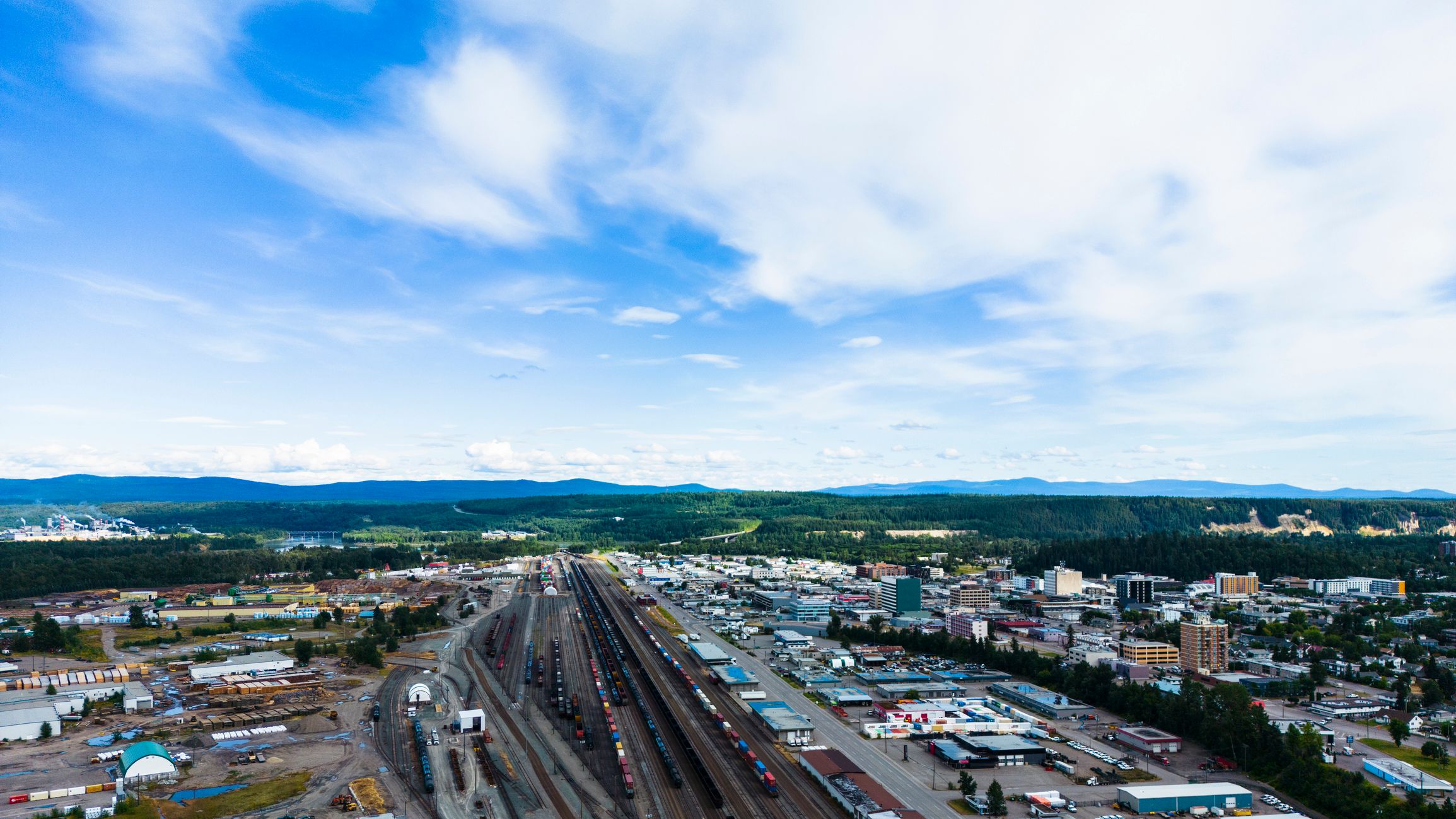 Aerial view of a Prince George, BC, showing a railway yard, industrial area, and forested hills under a blue sky.