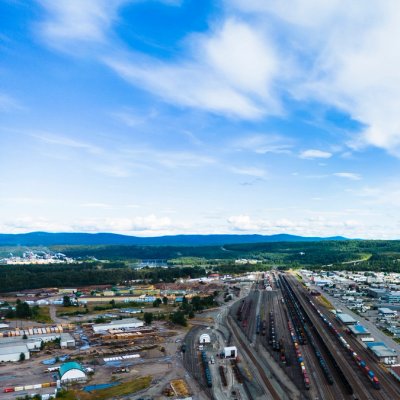 Aerial view of a Prince George, BC, showing a railway yard, industrial area, and forested hills under a blue sky.