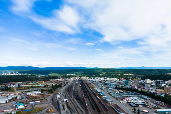 Aerial view of a Prince George, BC, showing a railway yard, industrial area, and forested hills under a blue sky.