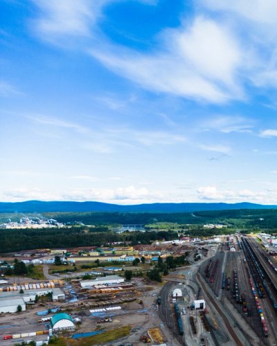 Aerial view of a Prince George, BC, showing a railway yard, industrial area, and forested hills under a blue sky.