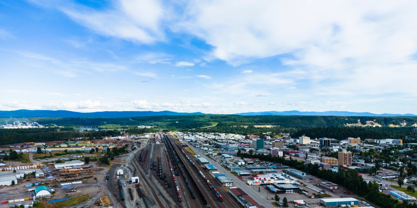 Aerial view of a Prince George, BC, showing a railway yard, industrial area, and forested hills under a blue sky.