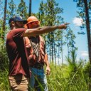 two men are standing in the middle of a forest pointing at something .