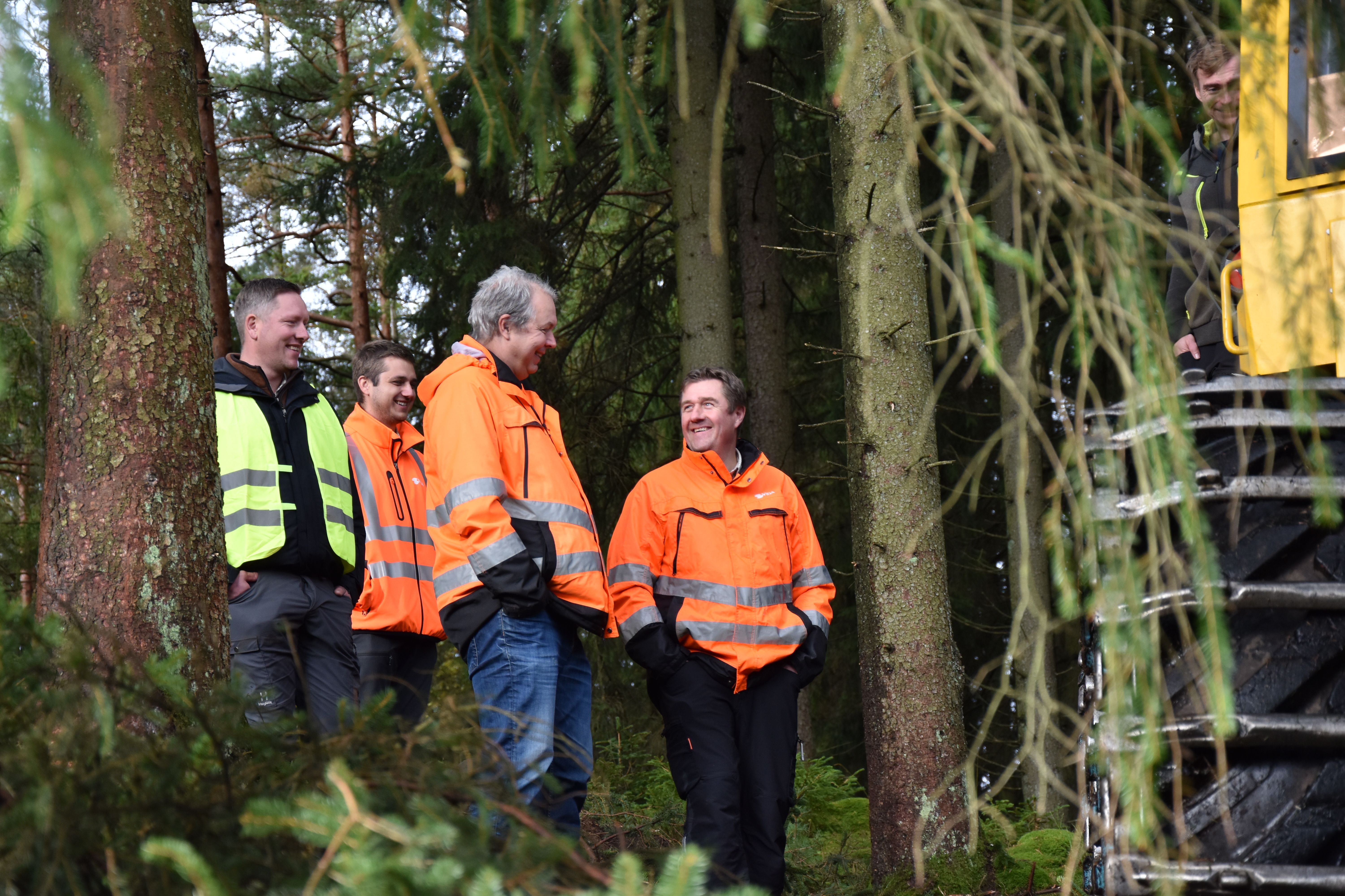 Five men in high-visibility jackets smiling and talking in a forest, next to a large yellow machine.