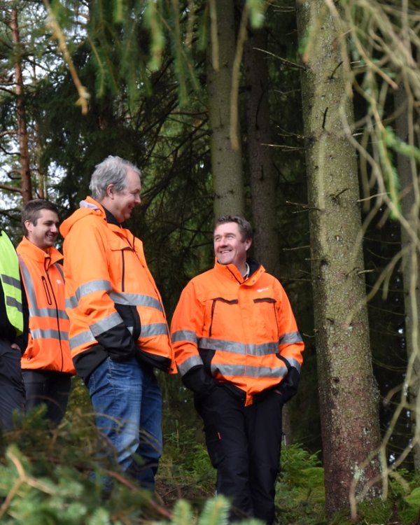 Five men in high-visibility jackets smiling and talking in a forest, next to a large yellow machine.