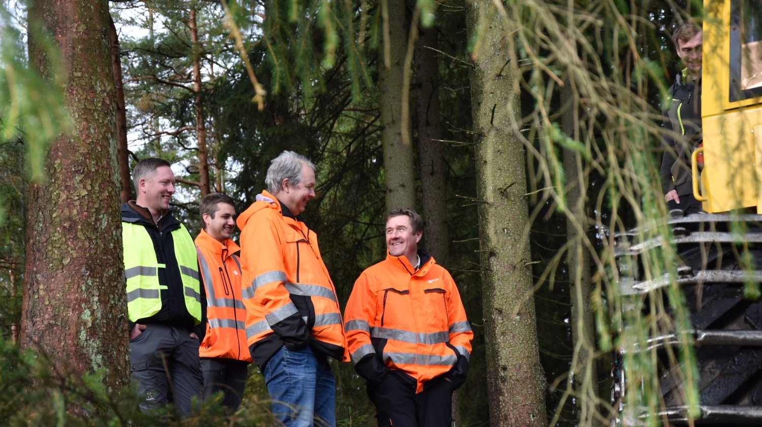 Five men in high-visibility jackets smiling and talking in a forest, next to a large yellow machine.