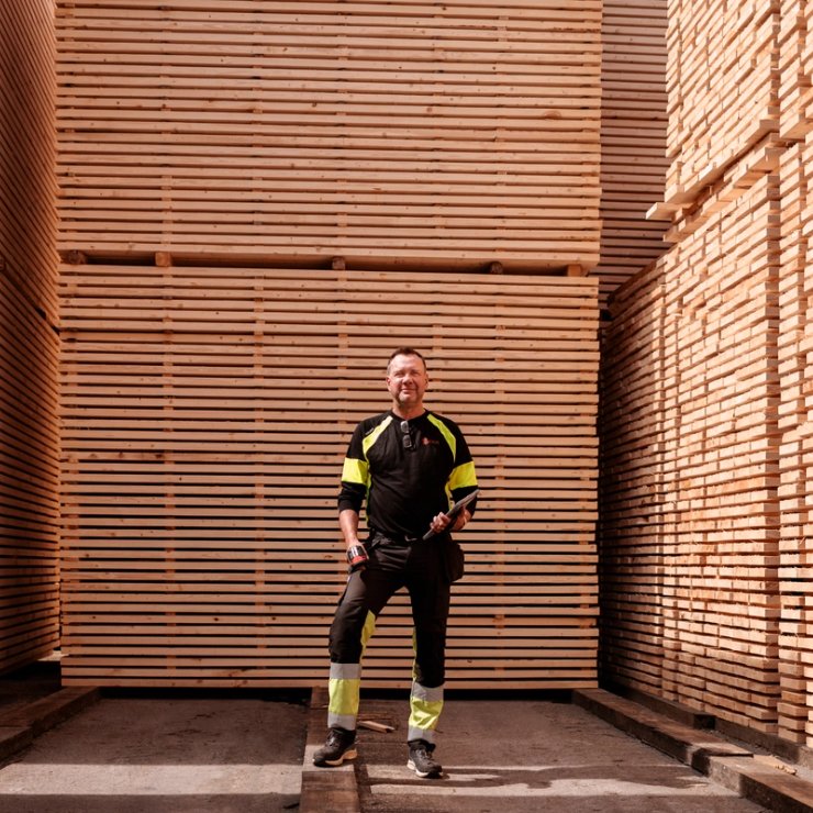 A man in work clothes holding a tablet stands among tall stacks of lumber.