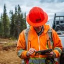 A man in an orange hard hat and high-visibility jacket uses a tablet at a logging site with stacked logs and heavy equipment in the background.