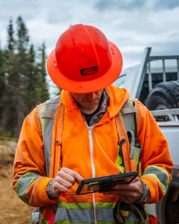 A man in an orange hard hat and high-visibility jacket uses a tablet at a logging site with stacked logs and heavy equipment in the background.