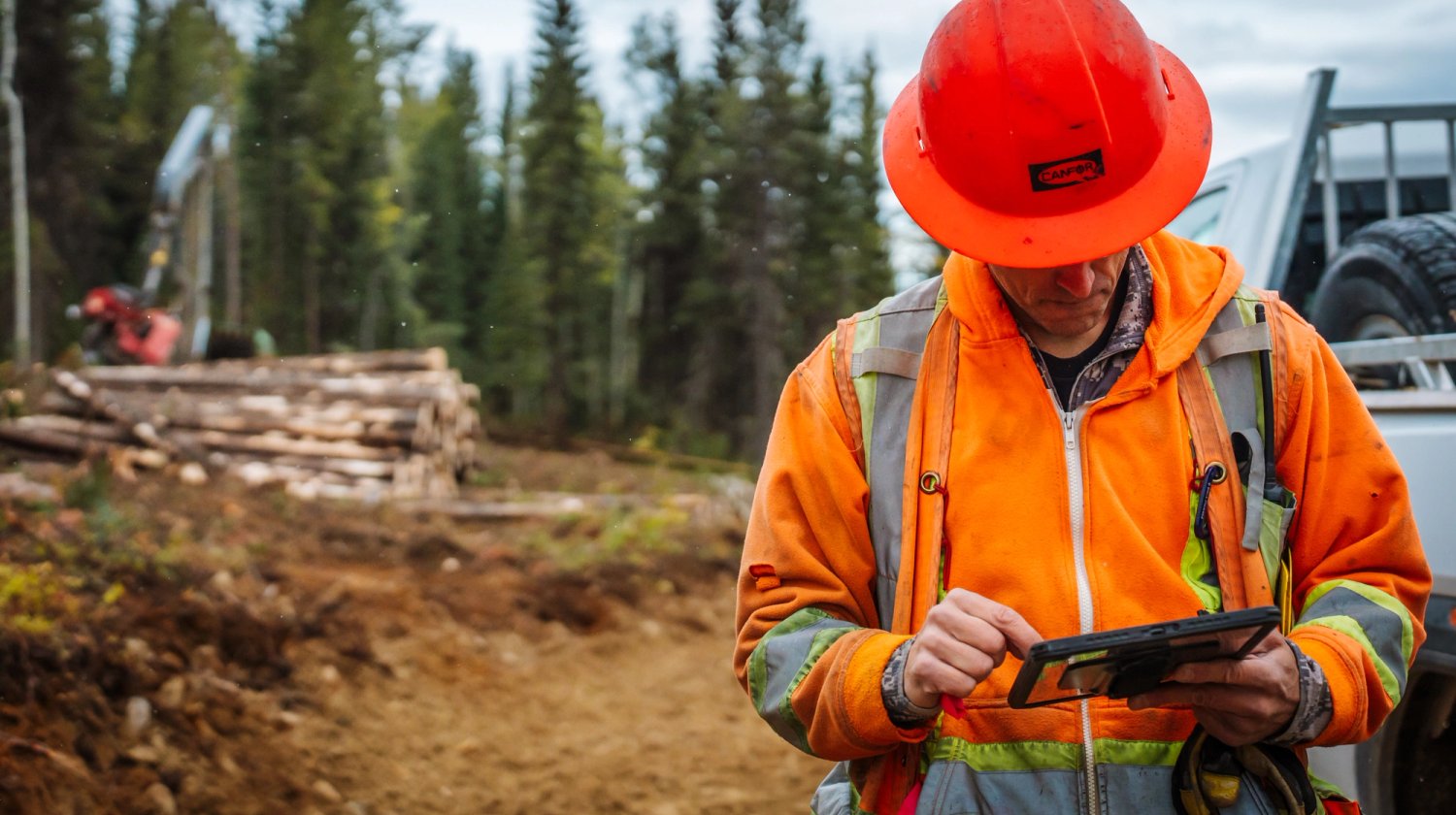 A man in an orange hard hat and high-visibility jacket uses a tablet at a logging site with stacked logs and heavy equipment in the background.