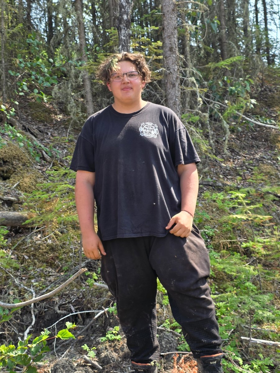 A young person with curly hair and glasses, wearing a black t-shirt and dark pants, stands in a forest.