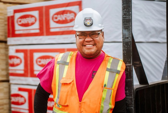 a man wearing a hard hat and safety vest is standing in front of a stack of Canfor lumber.