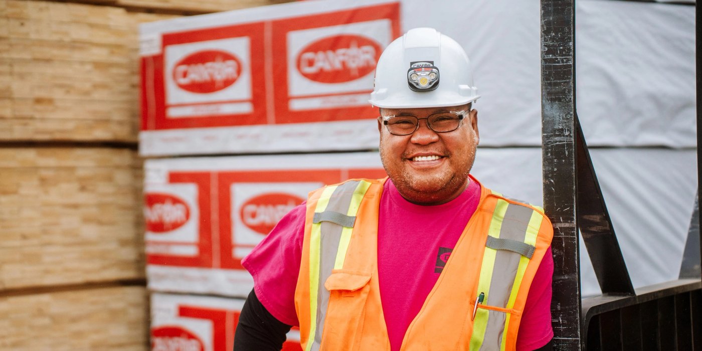 a man wearing a hard hat and safety vest is standing in front of a stack of Canfor lumber.