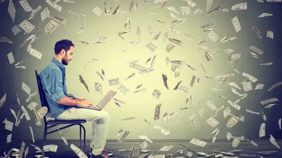 Young man using a laptop building online business making dollar bills cash falling down. Beginner IT entrepreneur under money rain. Success economy concept.jpeg
