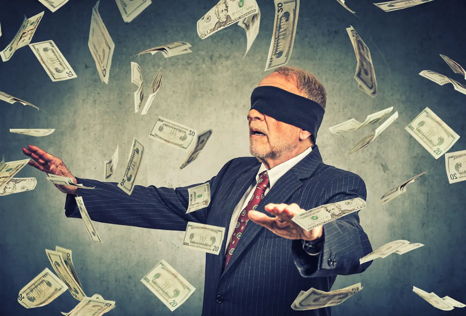 Blindfolded senior businessman trying to catch dollar bills banknotes flying in the air on gray wall background. Financial corporate success or crisis challenge concept-1.jpeg