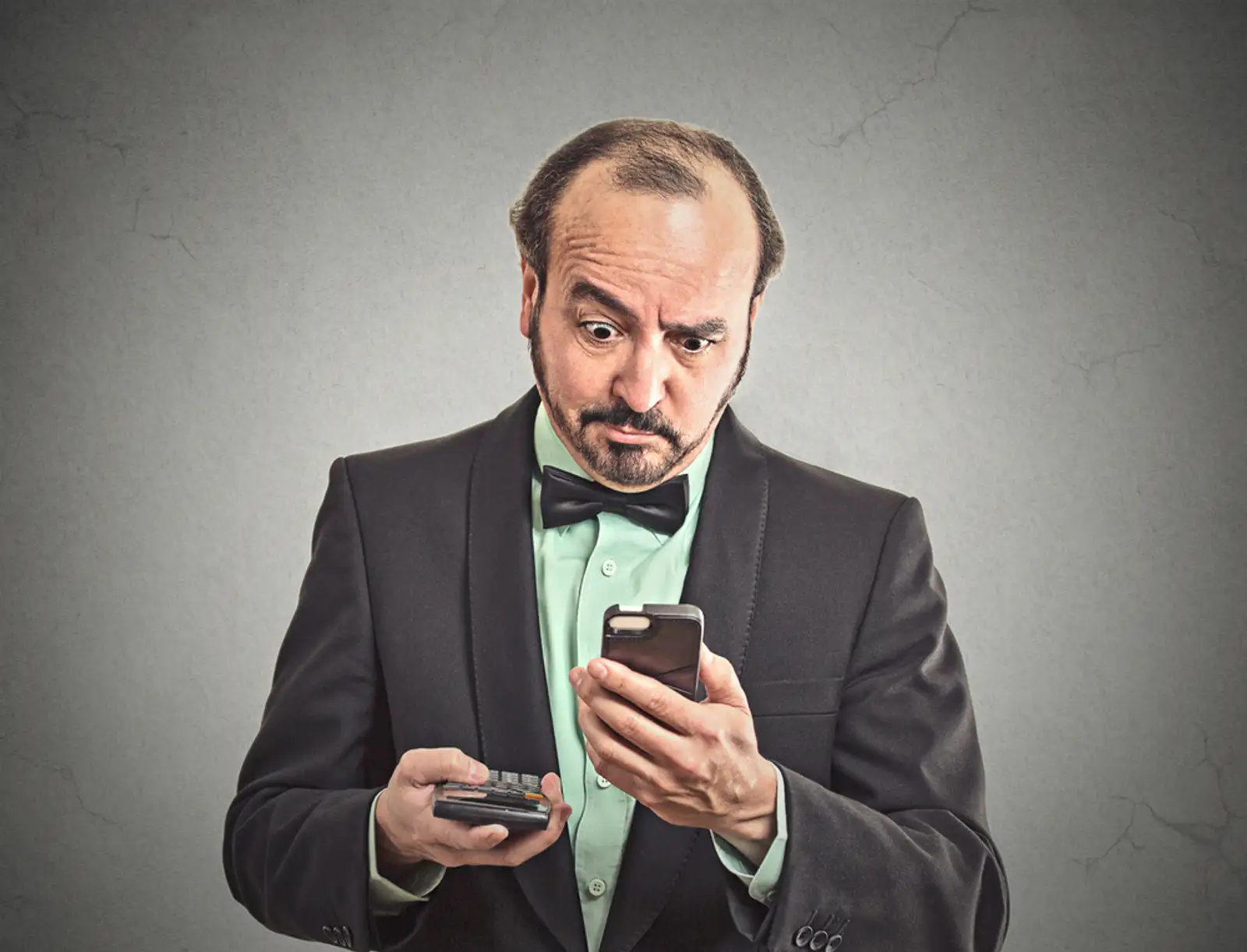 Portrait surprised businessman looking on smart phone holding calculator unexpected financial bills charges isolated grey wall background. Human face expression%2c emotion%2c body language%2c reaction.jpeg