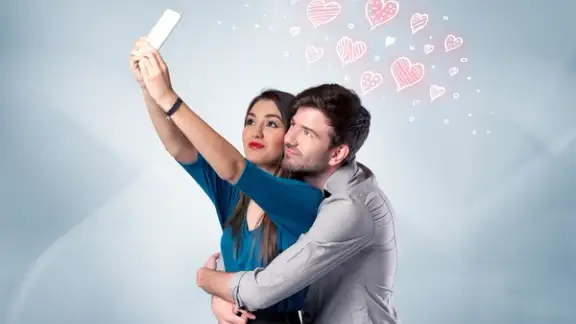 A young couple in love and drawn red hearts taking selfie with a mobile phone in the handsome guys hand in front of an empty clear grey wall background concept.jpeg