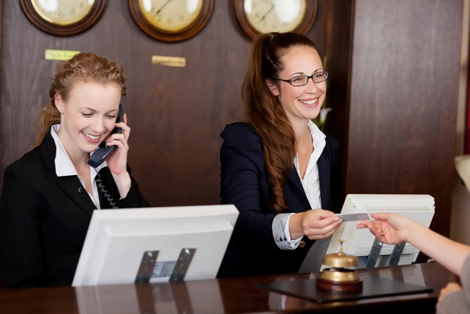Two beautiful young stylish receptionists at a reception desk%2c one talking on the telephone and the other handing a card to a customer.jpeg