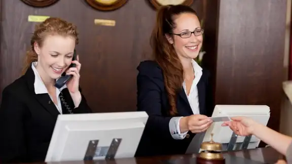 Two beautiful young stylish receptionists at a reception desk%2c one talking on the telephone and the other handing a card to a customer.jpeg