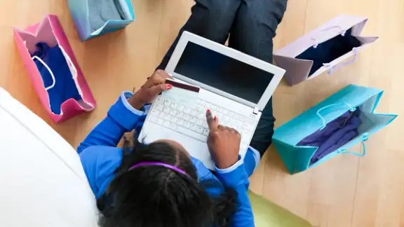 Pretty afro-american teenager using a laptop sitting between shopping bags on the floor in the living room