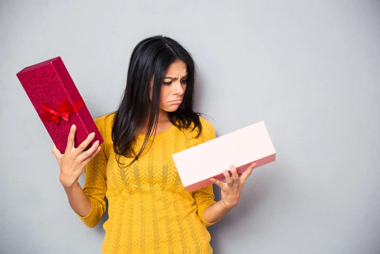 Unhappy young woman holding gift box over gray background.jpeg