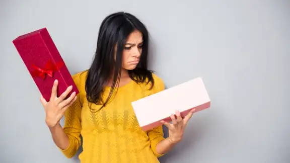 Unhappy young woman holding gift box over gray background.jpeg
