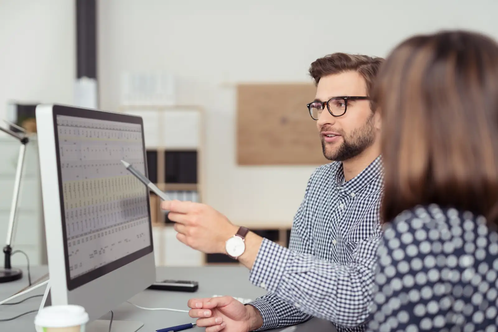 Proficient young male employee with eyeglasses and checkered shirt%2c explaining a business analysis displayed on the monitor of a desktop PC to his female colleague%2c in the interior of a modern office-1.jpeg