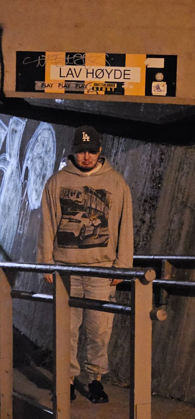 Isak standing in a dim underpass beneath a yellow low-clearance sign saying "Low height" in Norwegian, hands in his hoodie pockets.