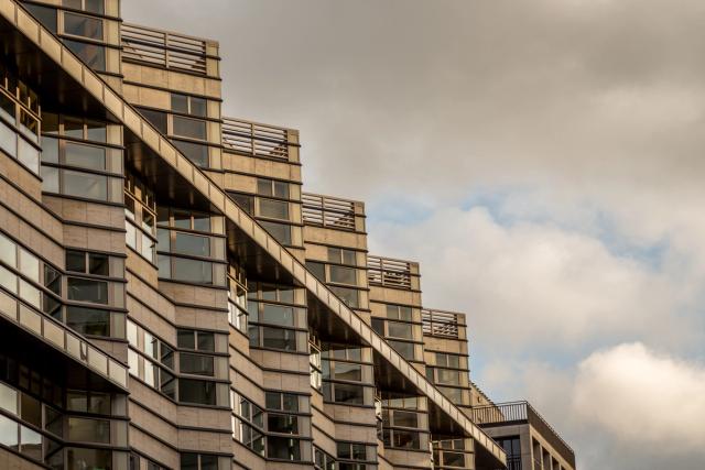 A stepped concrete apartment block seen from below, its balconied terraces receding diagonally into a bright, cloudy sky.