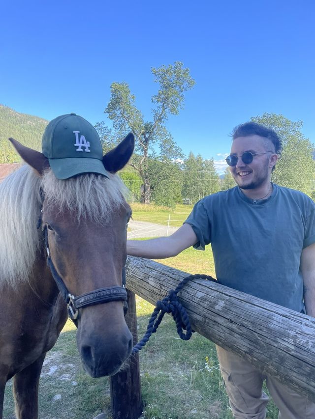Isak in sunglasses laughing beside a brown horse wearing his LA cap, green trees and mountains behind them.
