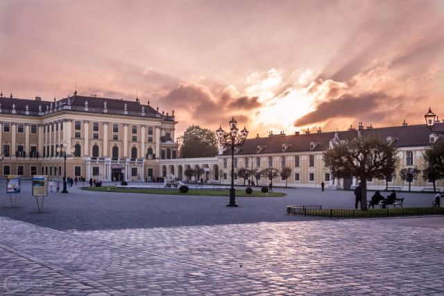 Sunset breaking pink and gold behind the long yellow wings of a baroque palace, in the foreground a wide cobbled forecourt is nearly empty.