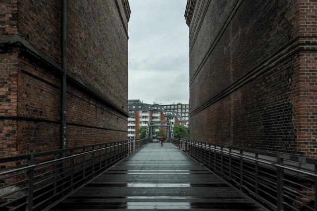 A wet steel footbridge running between two towering brick warehouse walls, a person in a red jacket at the far end.