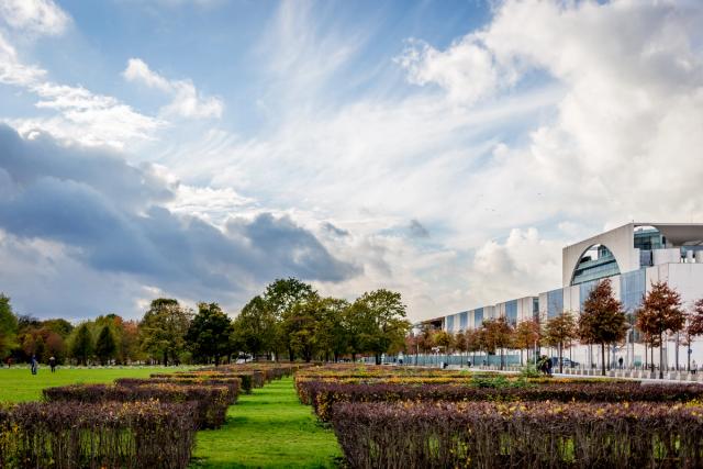 Clipped hedgerows lead across a lawn towards a curved glass-and-concrete government building, under a tall bank of cumulus.