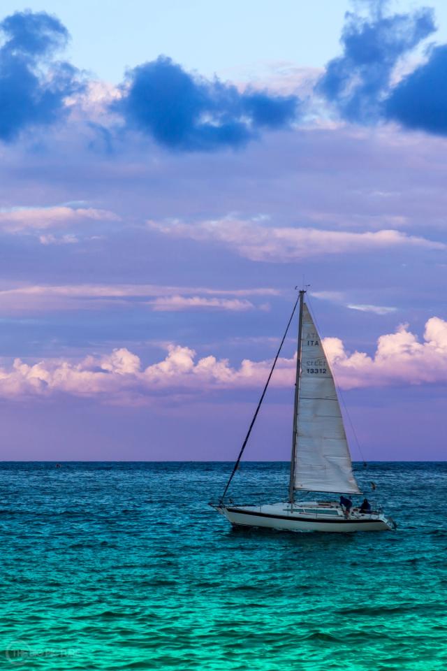 A single white sailboat under sail on turquoise water, beneath a wide purple and pink sky of scattered cloud.