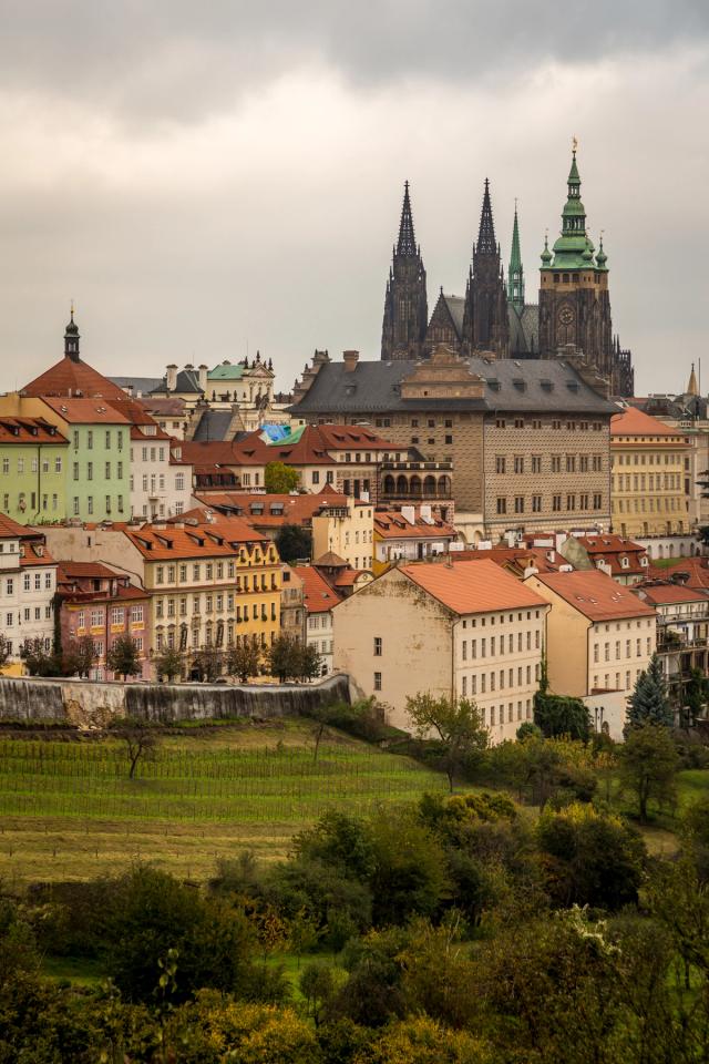 Terracotta rooftops climbing a hillside towards a gothic cathedral's spires and a green-domed tower, under flat grey cloud.