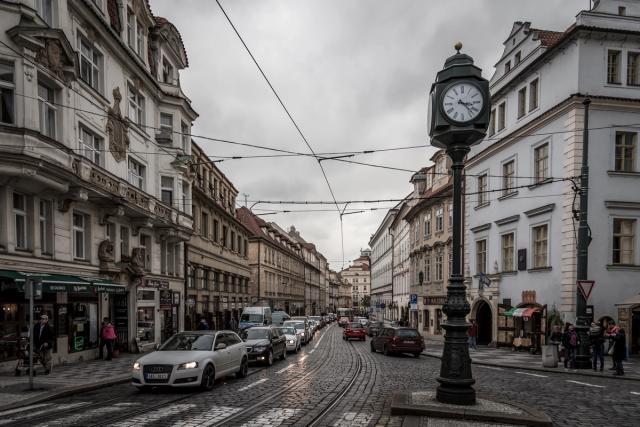 A cobbled street lined with pastel townhouses, tram wires overhead and an ornate black clock on a post in the foreground.