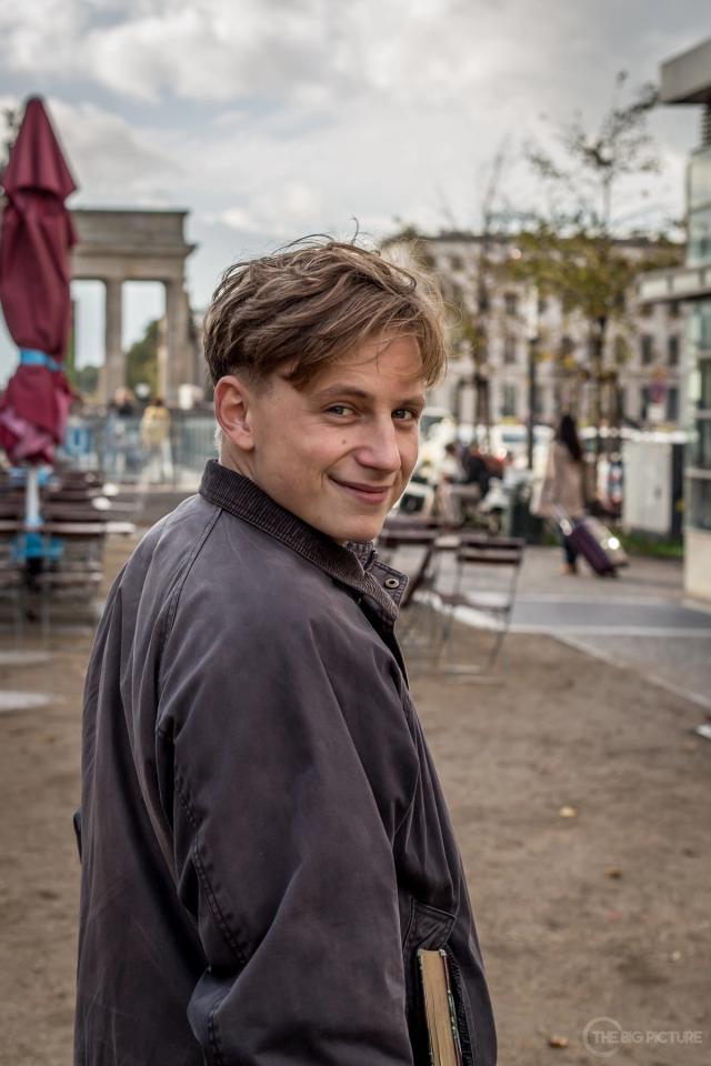A young man in a worn grey jacket glances back over his shoulder with a half smile, a book under his arm, a columned monument behind him.