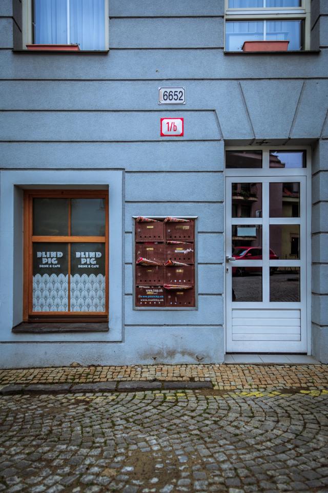 A pale blue building front on cobbles: a cafe window lettered Blind Pig, a bank of brown letterboxes, and a white glass door.