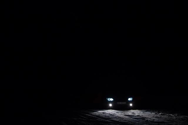 The headlights of a lone car cutting a small bright hole in total darkness on a snow-covered road.
