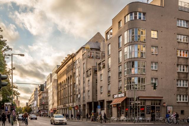 Evening light on a curved corner apartment block with a cafe at street level, traffic and cyclists on the wide road below.