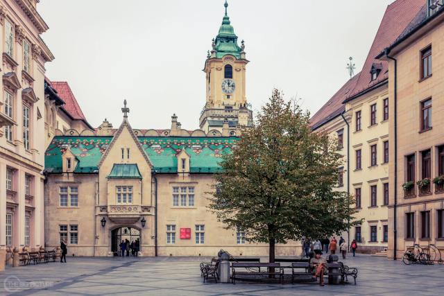 A near-empty old town square with a green-tiled roof and a clock tower, one person reading alone on a bench under a tree.