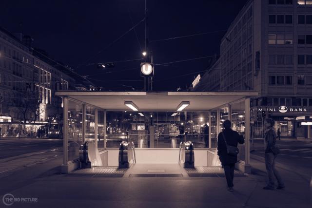 A lit glass underground entrance on an empty night square, escalators running down into it and two people passing by.