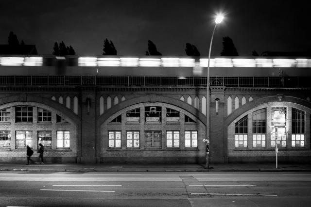 A train blurs across the top of a brick viaduct at night, in black and white, two people walking the empty pavement below.