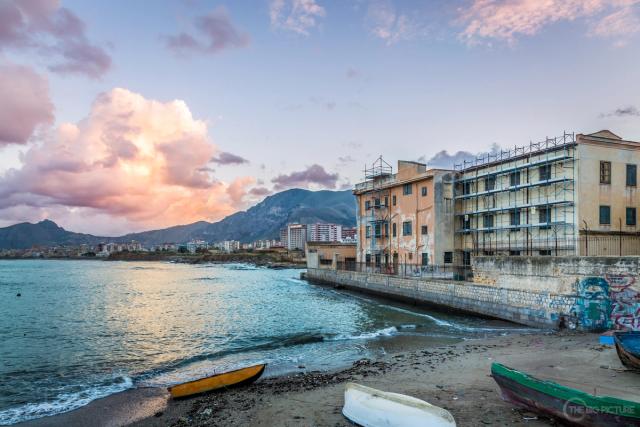 Small wooden boats pulled up on a grey beach at dusk, with faded seafront buildings and a mountain ridge beyond pink cloud.