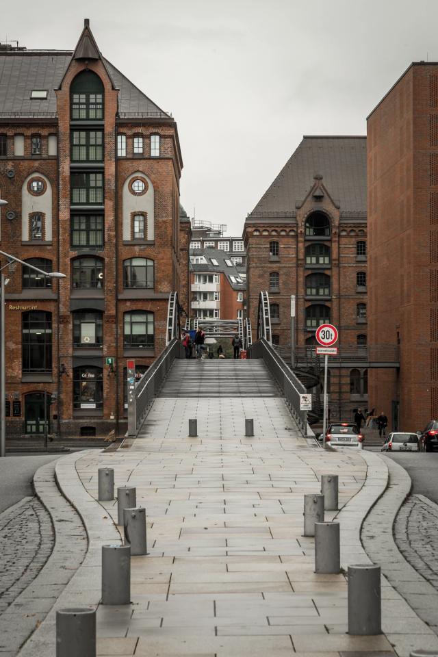 A wide paved ramp rising to a footbridge between red-brick warehouse blocks, bollards curving away in the foreground.