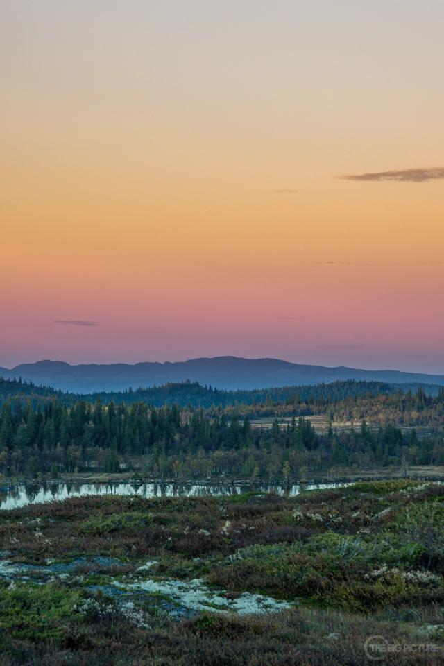 Layers of forested ridges under an orange and pink dusk sky, a thin lake catching the light below heather-covered ground.
