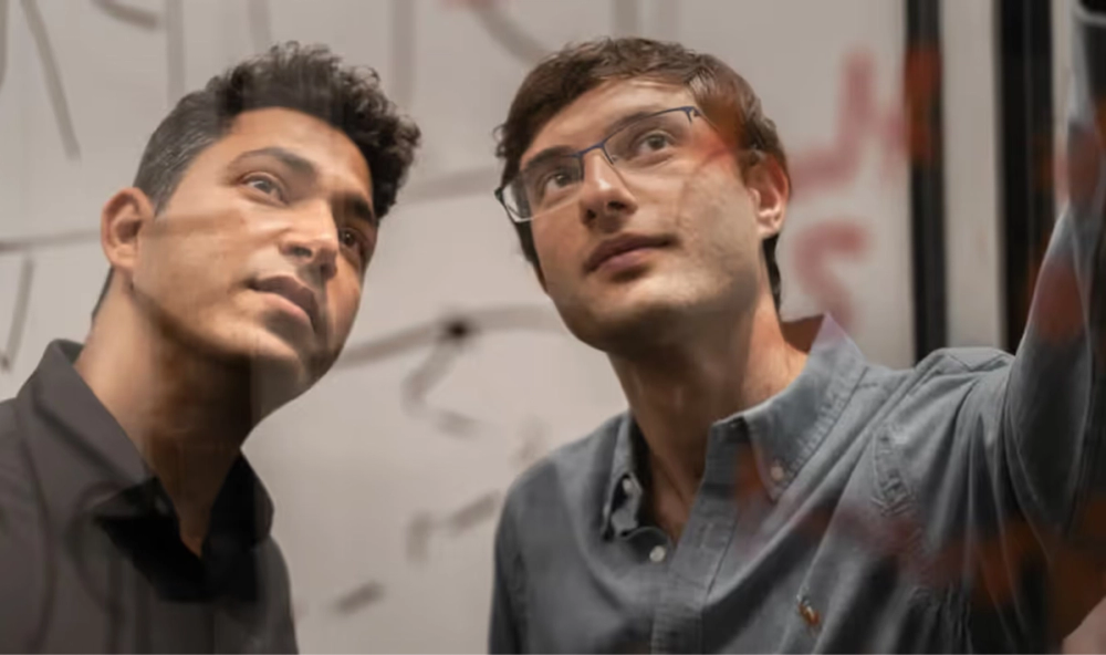 Two men looking intently at writing or drawings on a transparent board in a work or brainstorming session.