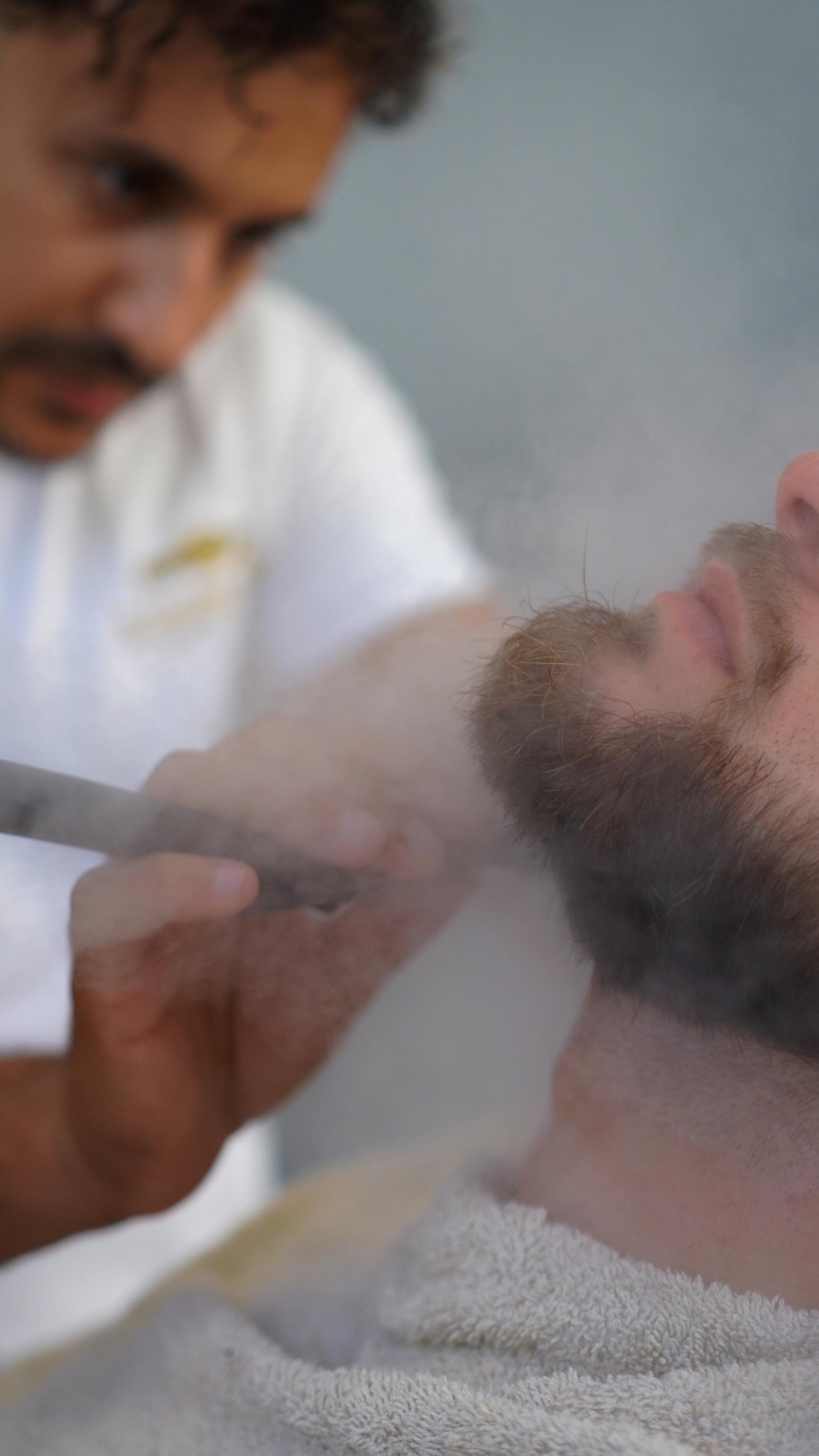 Barber shaving a client’s beard with a straight razor amid hot towel steam