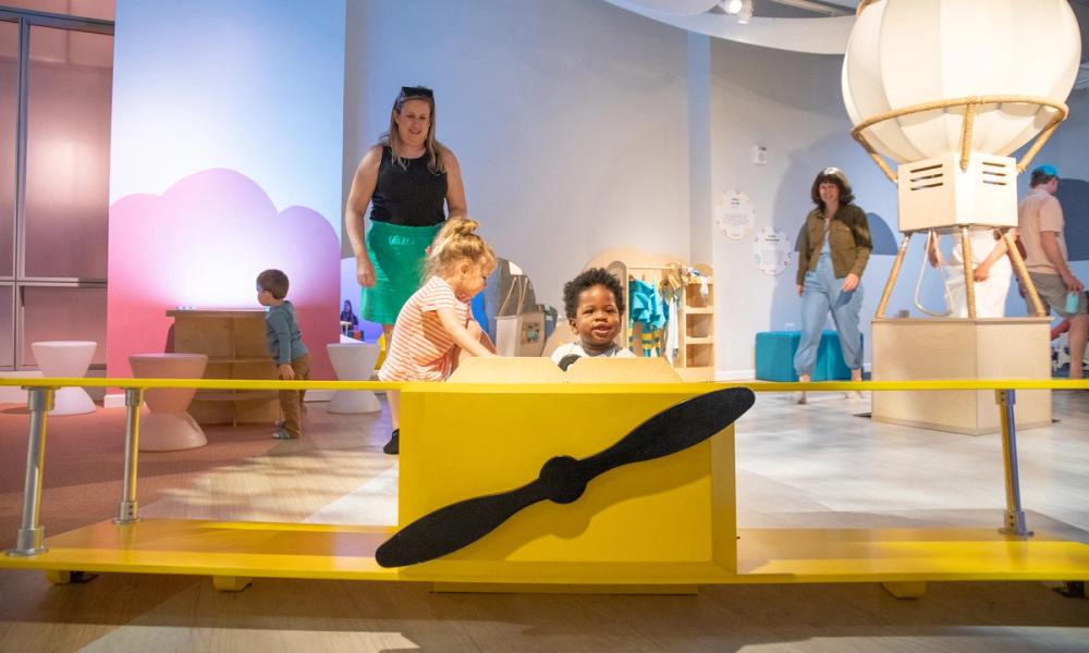 Two toddlers sit in a yellow wooden airplane as adults watch them.