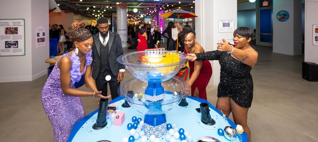 woman in purple sparkly dres, man in tux, women in red floor length dress, and woman in black sparkly jumpsuit playing at the fountain experience in placita magica
